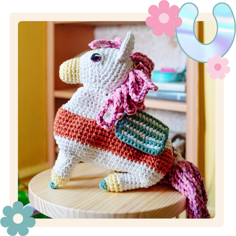 Crocheted unicorn toy with a pink mane and rainbow-patterned horn on a wooden surface.