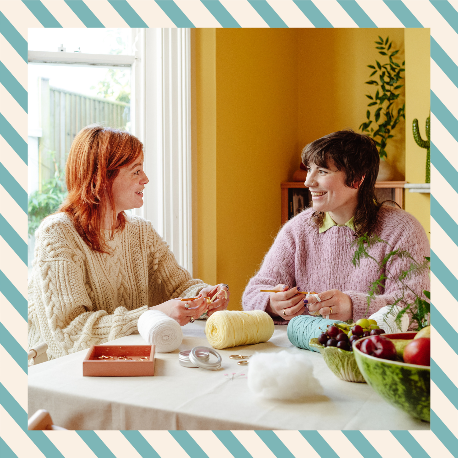 Two women sitting at a table with yarn and knitting materials, smiling and talking.